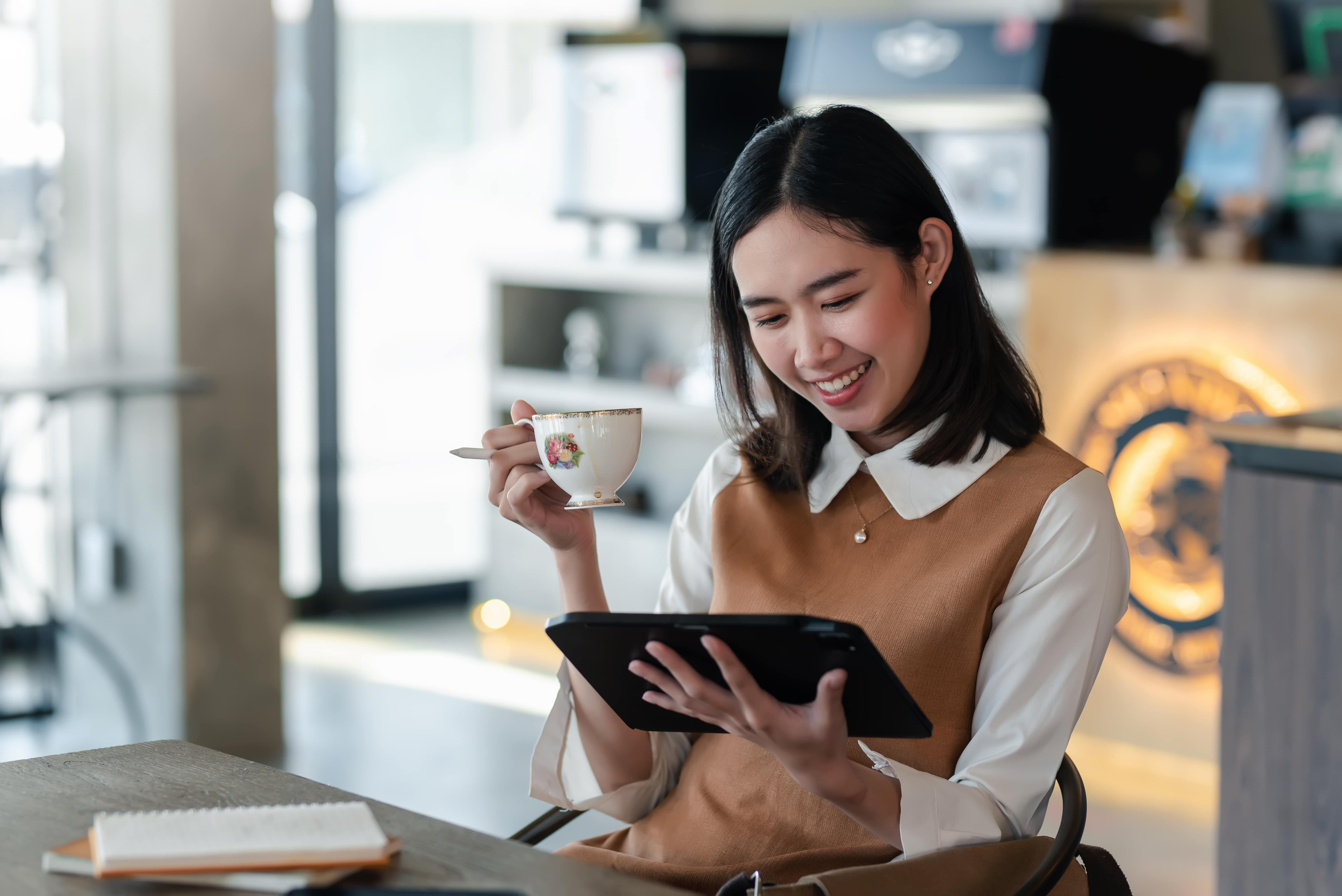 A woman sits in a café reading from a tablet and holding a tea cup.