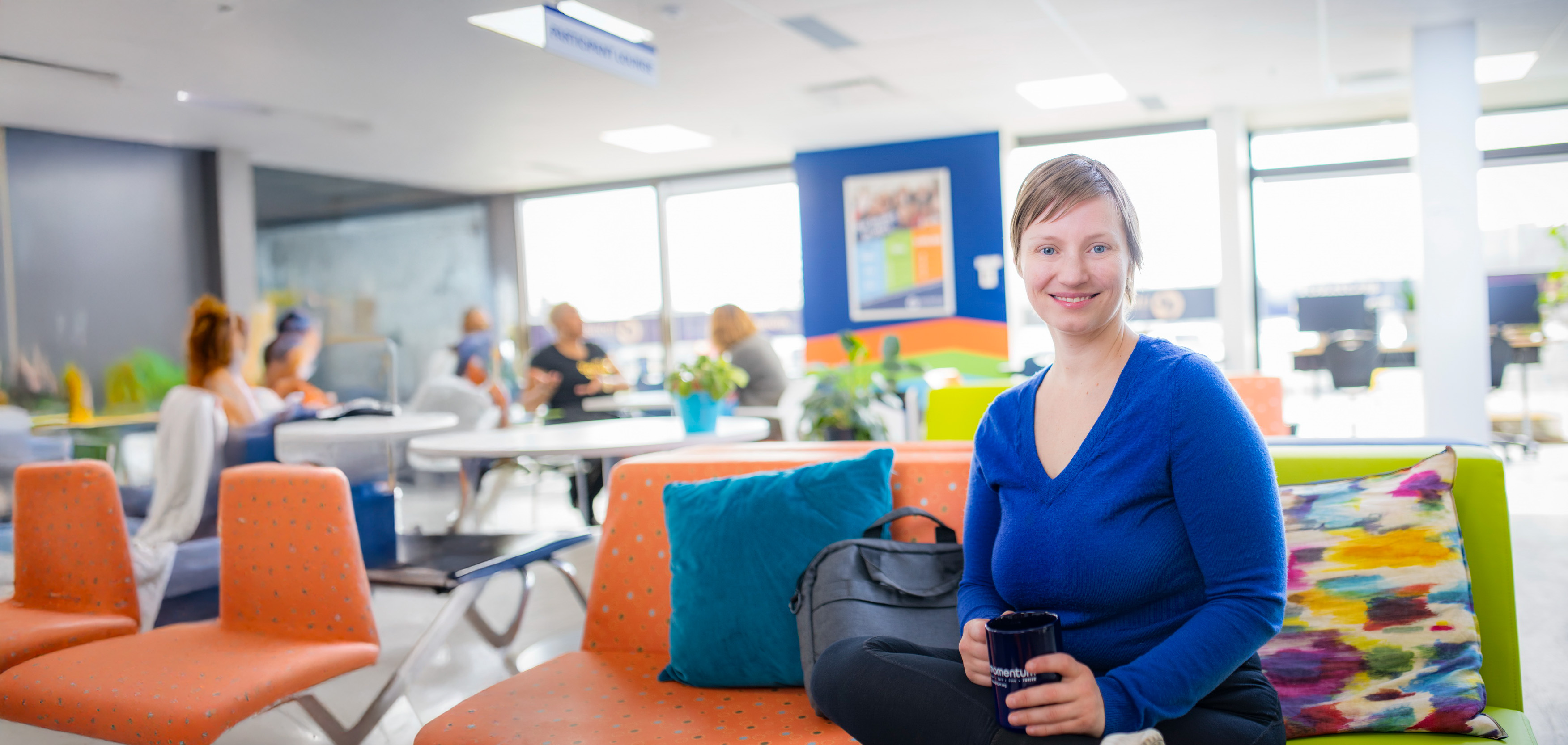 A woman sitting cross-legged on a couch in an office, with people sitting at tables in the background. She is smiling at the camera and holding a mug.